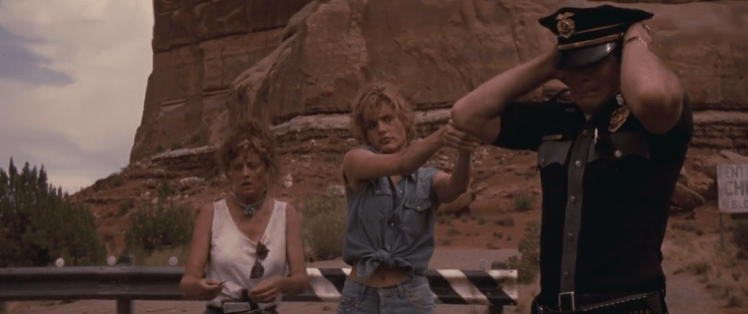 Thelma holds up the highway patrol cop, Louise stands behind her.